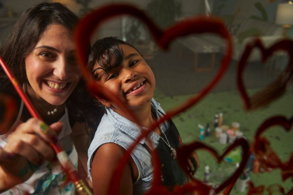 Young girl and mentor smiling into a mirror with painted hearts on it