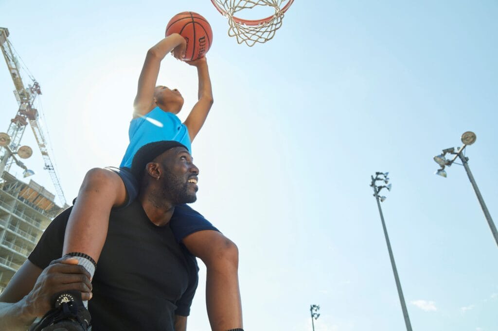 Young boy on mentor's shoulders reaching to put a basketball in the net